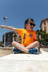Smiling child wearing sunglasses and an orange T-shirt sitting outdoors on a sunny day in Calpe,...