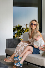 A stylish woman wearing sunglasses sits on a patio with a glass of wine while her young son plays...