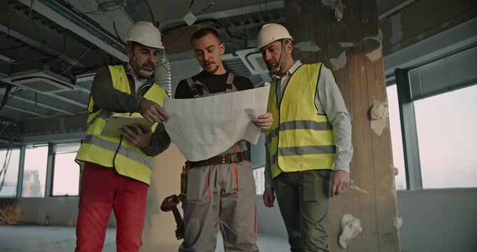 Engineers and a worker collaborate on a construction site, reviewing blueprints and discussing the project. They wear safety vests and hard hats.