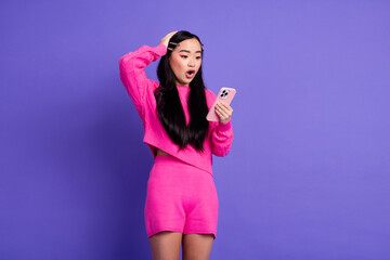 Stylish woman in pink attire astonished by smartphone message, standing against vibrant purple...