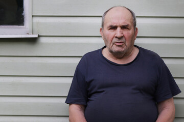 An old man in a blue T-shirt looks at the camera. A man after 80 years sits by the wall of the...