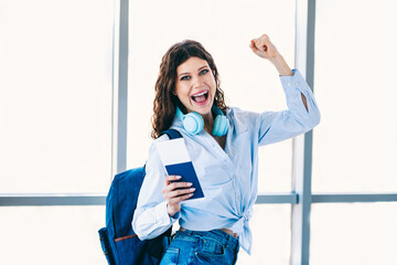 Excited young woman celebrating with a passport and headphones at the airport, ready for her travel...