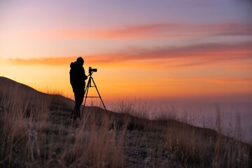 Silhouette of a photographer capturing a beautiful sunset from atop a grassy hillside with a camera on a tripod