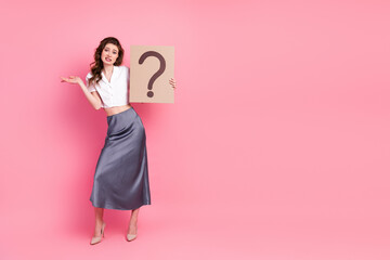 Young woman in stylish casual outfit holding a large question mark sign on bright pink background,...