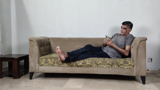 Man Sitting With Legs Stretched on Sofa Smiles Mischievously While Looking at Phone Reflecting Private Humor
