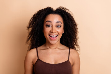 Joyful young woman with curly black hair in a brown top against a beige background expressing...