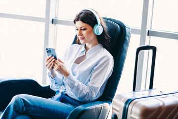 Young woman in headphones relaxing in airport lounge with smartphone and suitcase, enjoying leisure...