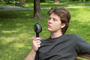 Handsome young caucasian man with hand electric fan suffering from heat at park. Heat Summer....
