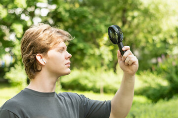 Handsome young caucasian man listen music and hand electric fan suffering from heat at park....