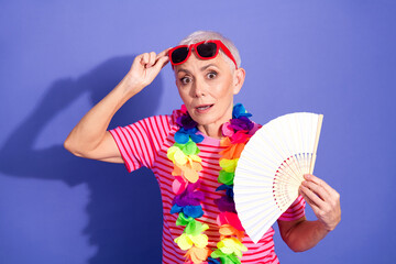 Playful elderly woman with vibrant lei and fan against a purple backdrop, showcasing fun, summer...