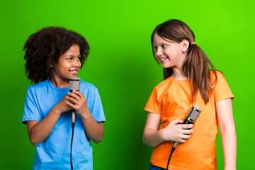Two cheerful young girls singing into microphones against a green background, sharing smiles and...