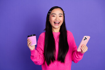 Joyful young woman holding a phone and coffee cup in a pink outfit against a vibrant purple backdrop