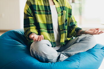 Young woman meditating at home in comfortable casual style, seated on beanbag, embracing peaceful...