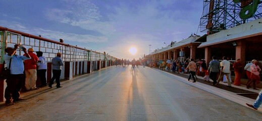Devotees walk toward the sacred Palani Murugan Temple during a serene sunset, casting long shadows...