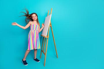 Happy schoolgirl posing near an easel wearing colorful summer dress against a turquoise background in joyful expression