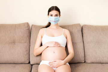 Pregnant woman in protective medical mask with coronavirus word is sitting on the sofa at home...