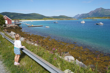 Little girl enjoying the view of the turquoise water harbour near Tromso with the fishing boats and red wooden shed on the foreground and mountains on the background.