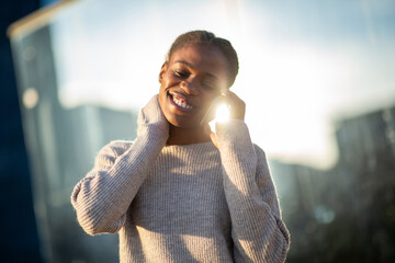 Young African woman smiling on phone call in sunlight with cozy sweater