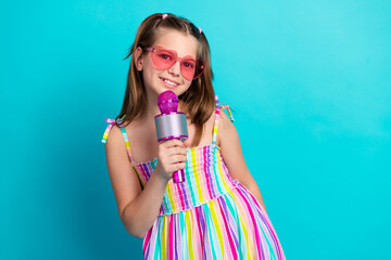 Charming young girl holding microphone wearing colorful striped dress and heart-shaped sunglasses...