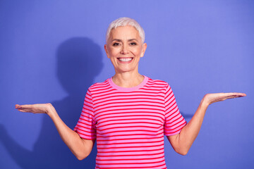 Portrait of a cheerful mature woman in a striped t-shirt gesturing with open hands against a purple...