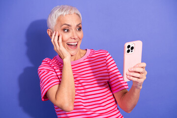 Excited Senior Woman Holding a Smartphone While Smiling Against a Vibrant Purple Background in...