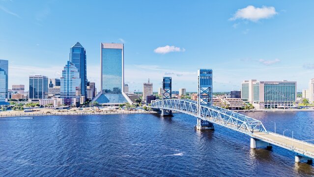 Aerial view of the bright blue bridge stretching over the dark river to the modern skyline under a sunny sky, Jacksonville, Florida, United States.
