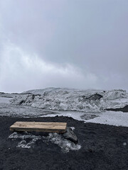 Wooden bench placed on black volcanic sand with snow-covered mountains in the background, under a...