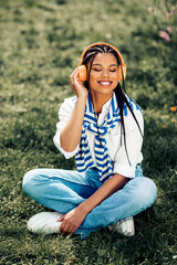 Charming woman with headphones sits outdoors enjoying music in the park during a sunny spring day,...