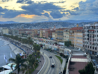 Scenic coastal view featuring palm trees lining a promenade, with colorful buildings and a winding road beside the ocean under a dramatic sky at sunset, capturing urban tranquility
