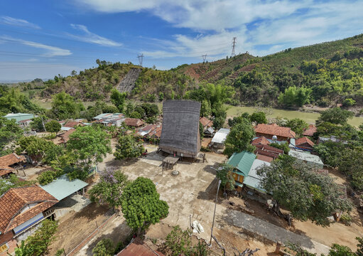 Aerial view of a traditional communal house (Nh&Atilde;&nbsp; R&Atilde;&acute;ng) standing tall amidst a village, set against rolling hills under a bright sky, Kon Tum, Kon Tum, Vietnam.