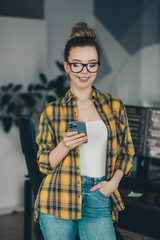 Qualified professional woman with glasses using smartphone in creative workspace, wearing casual...