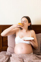 Hungry pregnant woman relaxing in bed is eating greedily a piece of cake and has a dirty mouth....