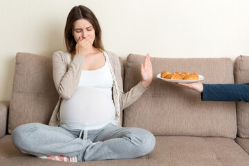 Pregnant woman relaxing on the sofa makes stop gesture to croissants. Expecting mother refuses to...
