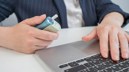 Business woman smoking an electronic cigarette while working on a laptop. Tobacco heating system. 