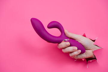 Female hand sticking out with raspberry sex toy from pink paper background.