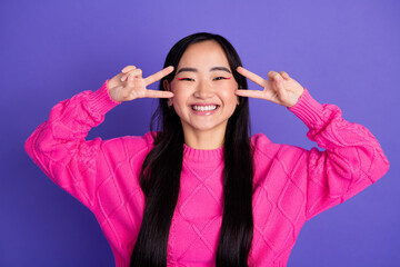 Cheerful young woman in vibrant pink sweater posing with peace gesture on purple background