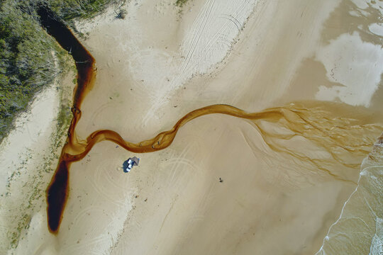 Aerial view of a dark river snaking its way across a sandy beach to meet the light-colored ocean, creating a stark contrast of colors, Saxmundham, England, United Kingdom.