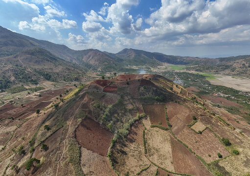 Aerial view of a volcano covered with agricultural fields, the terrain's reddish-brown hues juxtaposed against the sky's blue and white, Chu Pah, Gia Lai, Vietnam.
