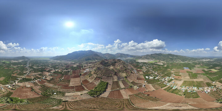 Aerial view of earth-toned fields juxtapose with lush green patches, nestled beneath rolling mountains against a clear blue sky speckled with clouds, Chu Pah, Gia Lai, Vietnam.