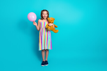 Smiling girl in colorful dress holding pink balloon and teddy bear against teal backdrop...