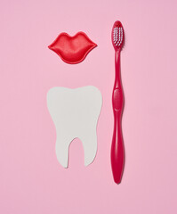 Red toothbrush, red decorative lips and paper cutout white tooth placed on pink background.