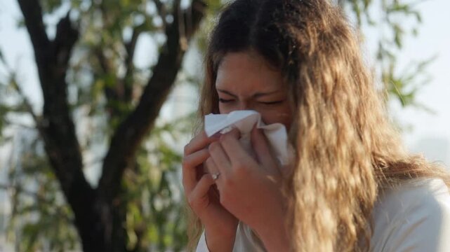 Portrait of young Asian woman sneezes and stands in green spring park closeup. Female with loose hair shows allergy symptoms and wipes nose with tissues spending time in nature