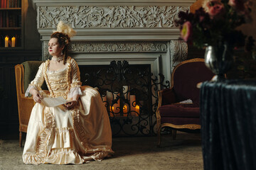 Caucasian young adult woman wearing elaborate Baroque style gown sitting on chair holding sheet of...