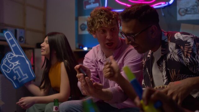 Side view shot of Caucasian man as sports fan watching match on TV and pointing at phone making bets while cheering with multiethnic group of friends, copy space