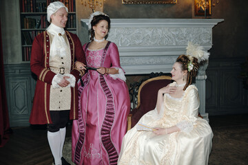 Three Caucasian adults dressed in Baroque period costumes interacting in ornate Renaissance style...