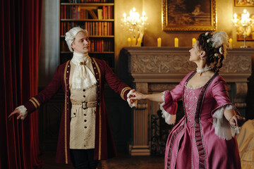 Young man and woman dressed in Baroque period costumes performing elegant dance in ornate...