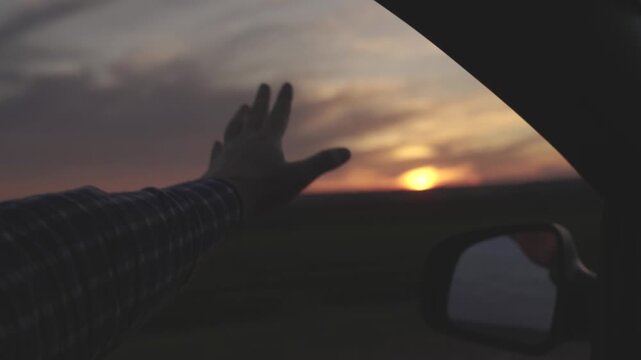 The hand from the car catches the wind against the backdrop of the setting sun, long-distance travel, free movement by car on a long journey, the road of rest and entertainment.
