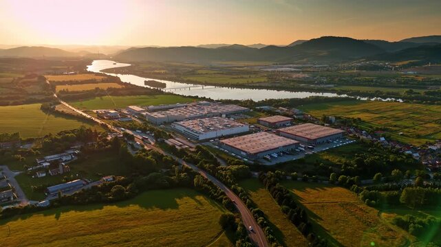 Flying over the rural area near the river at sunset time. Cars move by the road along the large industrial premises. Slovakia. Aerial view.