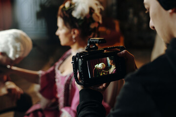 Caucasian young adult woman wearing Baroque style costume sitting while being filmed by young adult Caucasian man holding professional video camera during historical reenactment