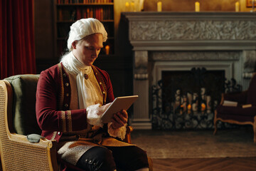 Caucasian young adult man with powdered wig and period costume sitting in ornate chair using...